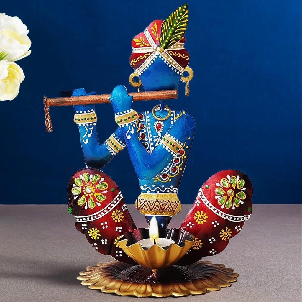 A decorative painted figurine of a seated blue flute-player with red patterned legs and a gold lotus base, styled on a tabletop with white flowers and a dark blue backdrop.