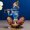 A decorative painted figurine of a seated blue flute-player with red patterned legs and a gold lotus base, styled on a tabletop with white flowers and a dark blue backdrop.